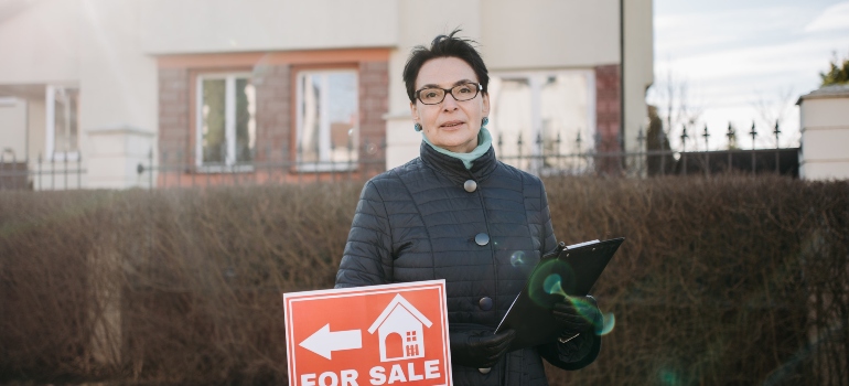 une femme debout devant une maison où est affichée une pancarte « À vendre »